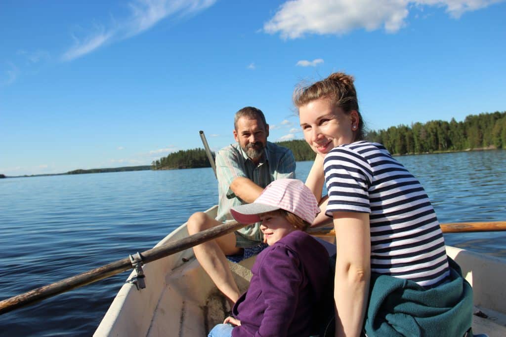 Kesäinen veneretki. Mies soutaa, nainen ja hymyilevä lapsi veneessä kuvan etualalla. Aholan mökit Padasjoki.